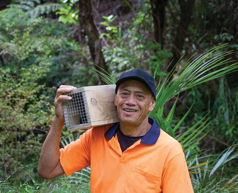 Man wearing a cap and orange shirt standing in the bush with a box pest trap on his shoulder.