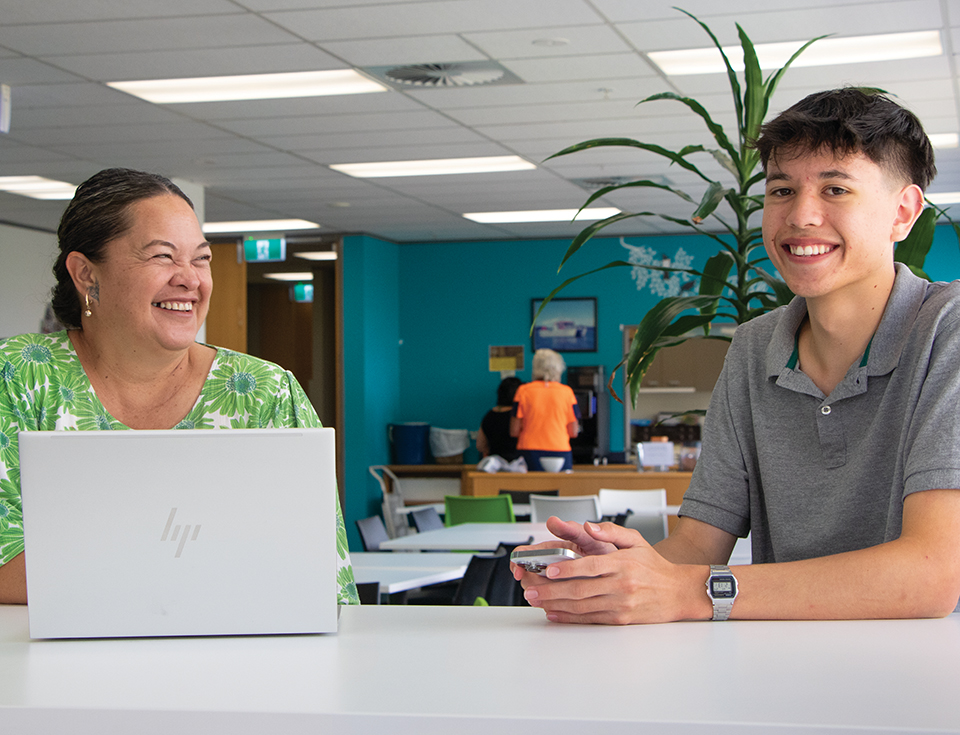 Woman with laptop and young man at a table smiling with pot plant in the background.